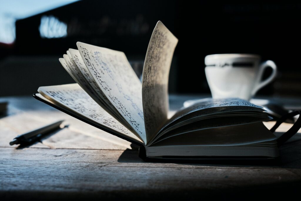 Photo of a notebook on a desk, its pages loosely open and full of writing. Beside it is a pen, and behind it is a cup.